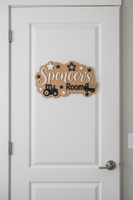 A scalloped wooden sign for a child's room featuring the text 'Spencer's Room' in white and navy blue, decorated with star cutouts and tractor and truck illustrations, displayed on a textured gray fabric background.