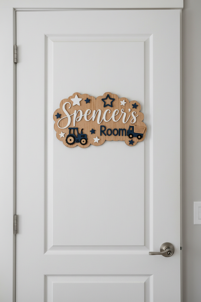 A scalloped wooden sign for a child's room featuring the text 'Spencer's Room' in white and navy blue, decorated with star cutouts and tractor and truck illustrations, displayed on a textured gray fabric background.