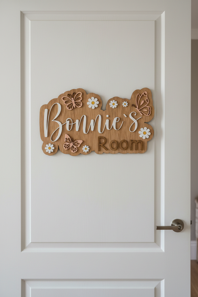 A custom wooden sign reading 'Bonnie's Room' with decorative cutout butterflies and white daisy flowers, displayed on a textured gray fabric background.