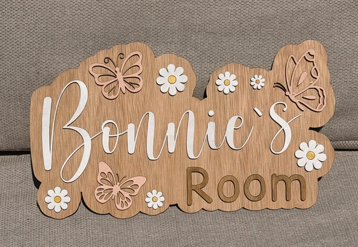 A custom wooden sign reading 'Bonnie's Room' with decorative cutout butterflies and white daisy flowers, displayed on a textured gray fabric background.