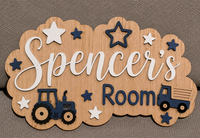 A scalloped wooden sign for a child's room featuring the text 'Spencer's Room' in white and navy blue, decorated with star cutouts and tractor and truck illustrations, displayed on a textured gray fabric background.