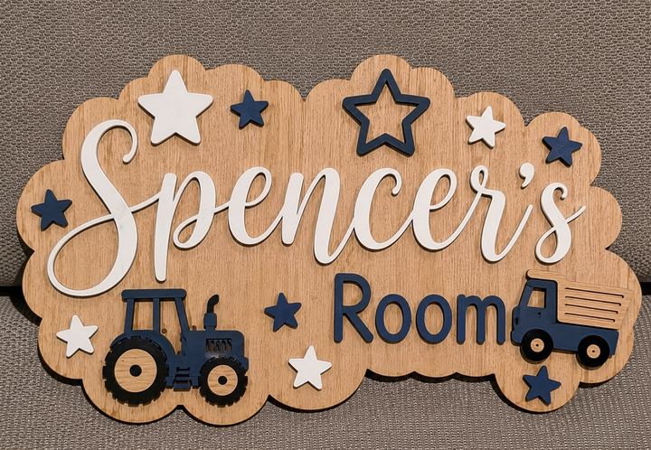 A scalloped wooden sign for a child's room featuring the text 'Spencer's Room' in white and navy blue, decorated with star cutouts and tractor and truck illustrations, displayed on a textured gray fabric background.