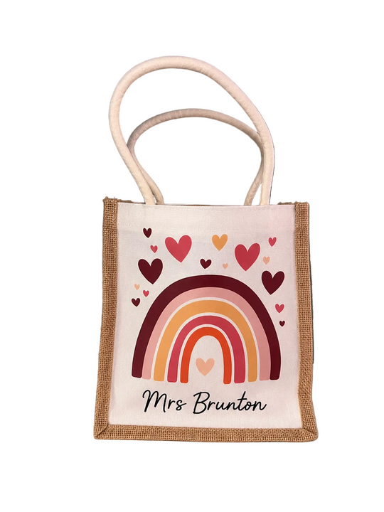 A small jute tote bag with a white printed panel featuring a rainbow, hearts, and the text 'Mrs Brunton', shown standing on a white surface against a dark gray wall.