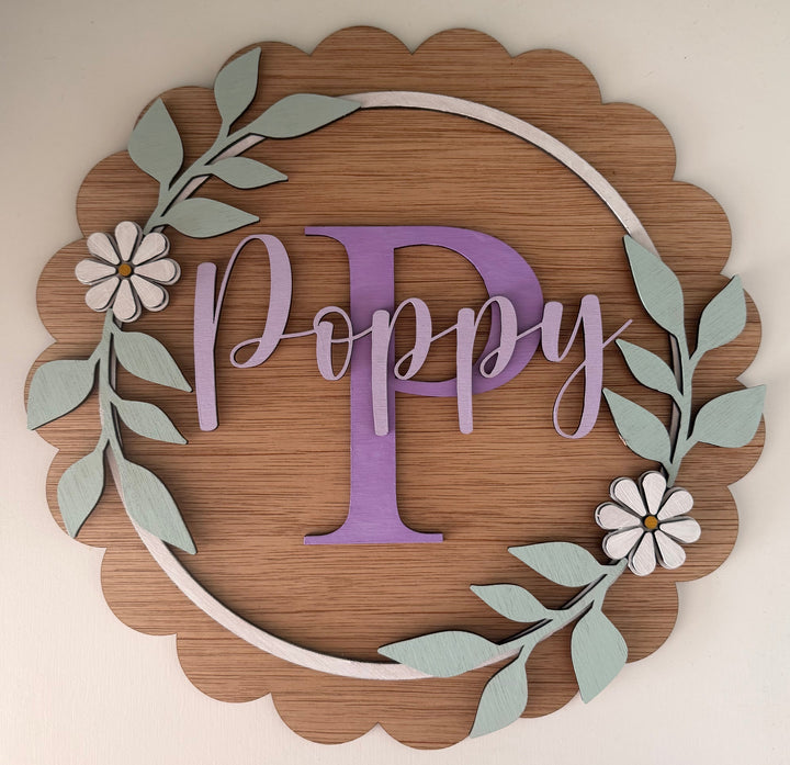 A personalized wooden wall sign with a scalloped oak-look border, a central lavender letter 'P', the name 'Poppy' in lavender script, a white circular ring, and pale green leafy branches with white flowers, shown mounted on a white wall.