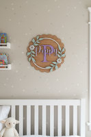 A personalized wooden wall sign with a scalloped oak-look border, a central lavender letter 'P', the name 'Poppy' in lavender script, a white circular ring, and pale green leafy branches with white flowers, shown mounted on a white wall.