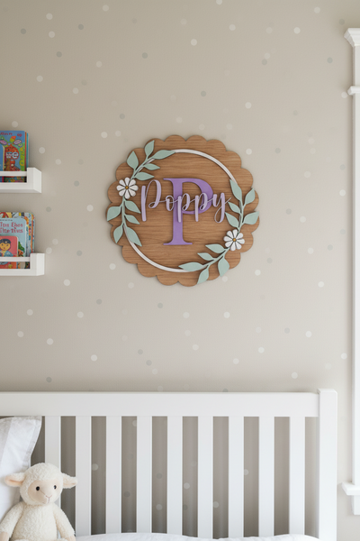 A personalized wooden wall sign with a scalloped oak-look border, a central lavender letter 'P', the name 'Poppy' in lavender script, a white circular ring, and pale green leafy branches with white flowers, shown mounted on a white wall.